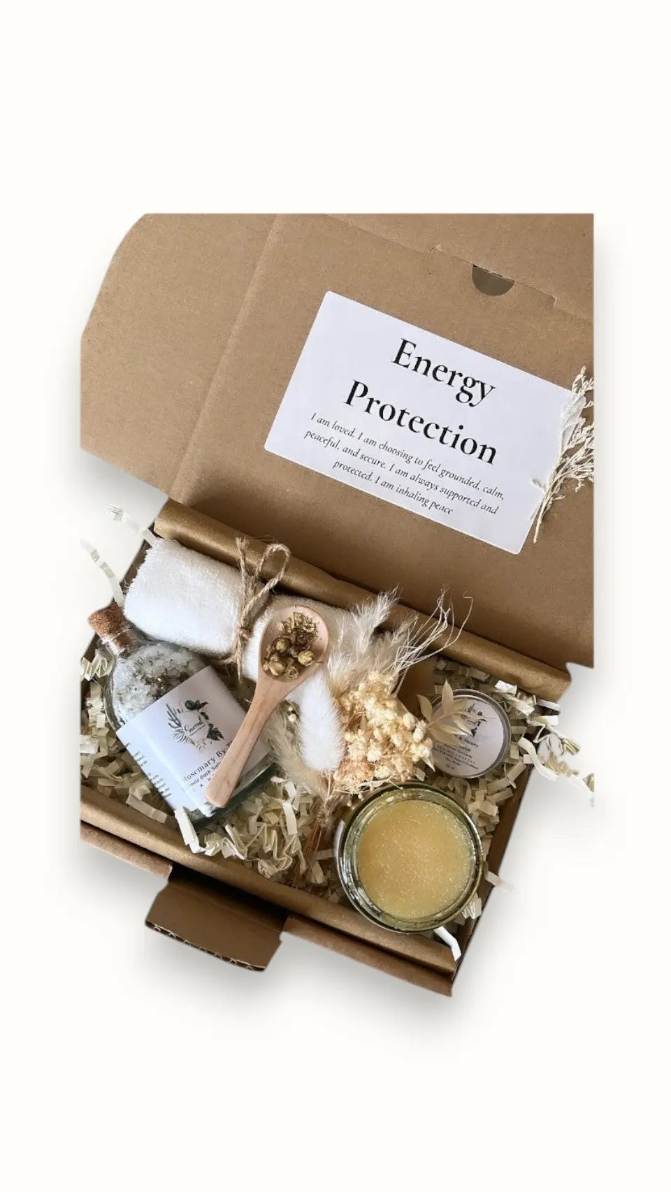 A gift box labeled 'Energy Protection' with various items including a bath salt, lip balm, sugar scrub, a dried flower, a bowl, a spoon, and a towel, all packaged with tissue paper and shredded paper for protection. Positive affirmation gifts, Christmas gift box and Spa gift set