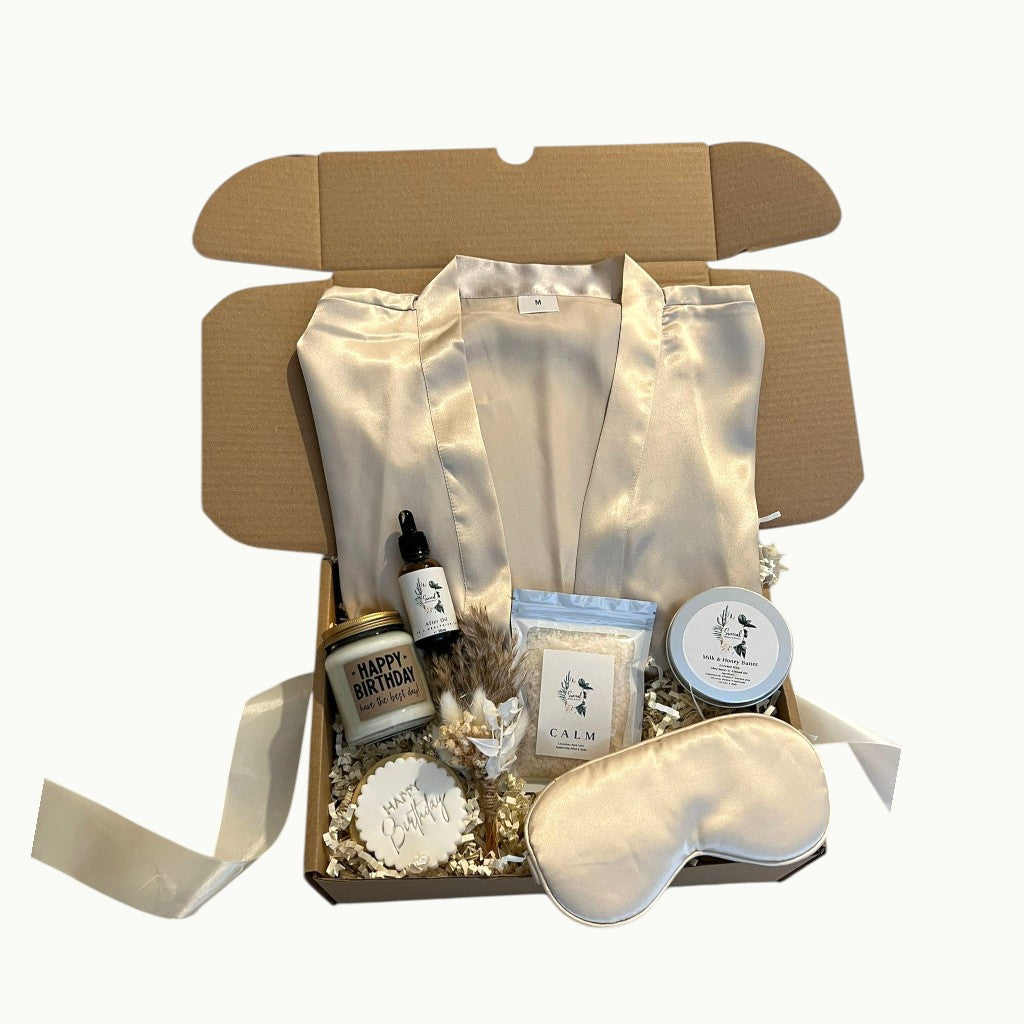 Self care gift box featuring a satin cream robe, silk eye mask, skincare products, bath salts, candle and personalised cookie, beautifully arranged in a pamper hamper