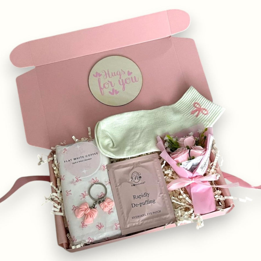 A gift box with various personal care items including a flat white coffee bar, a pair of pink socks with a floral design, a pink bow keyring, a packet of De Puff eye patches, a mini rose flower, and a coaster, all arranged on decorative tissue paper.
