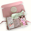 A gift box with various personal care items including a flat white coffee bar, a pair of pink socks with a floral design, a pink bow keyring, a packet of De Puff eye patches, a mini rose flower, and a coaster, all arranged on decorative tissue paper.