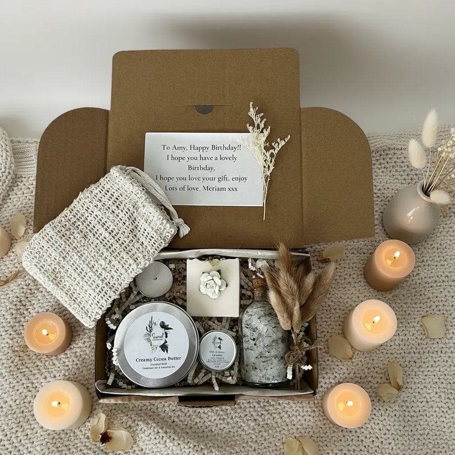Gift box with bath products and a card on a textured surface with candles and decorative elements.