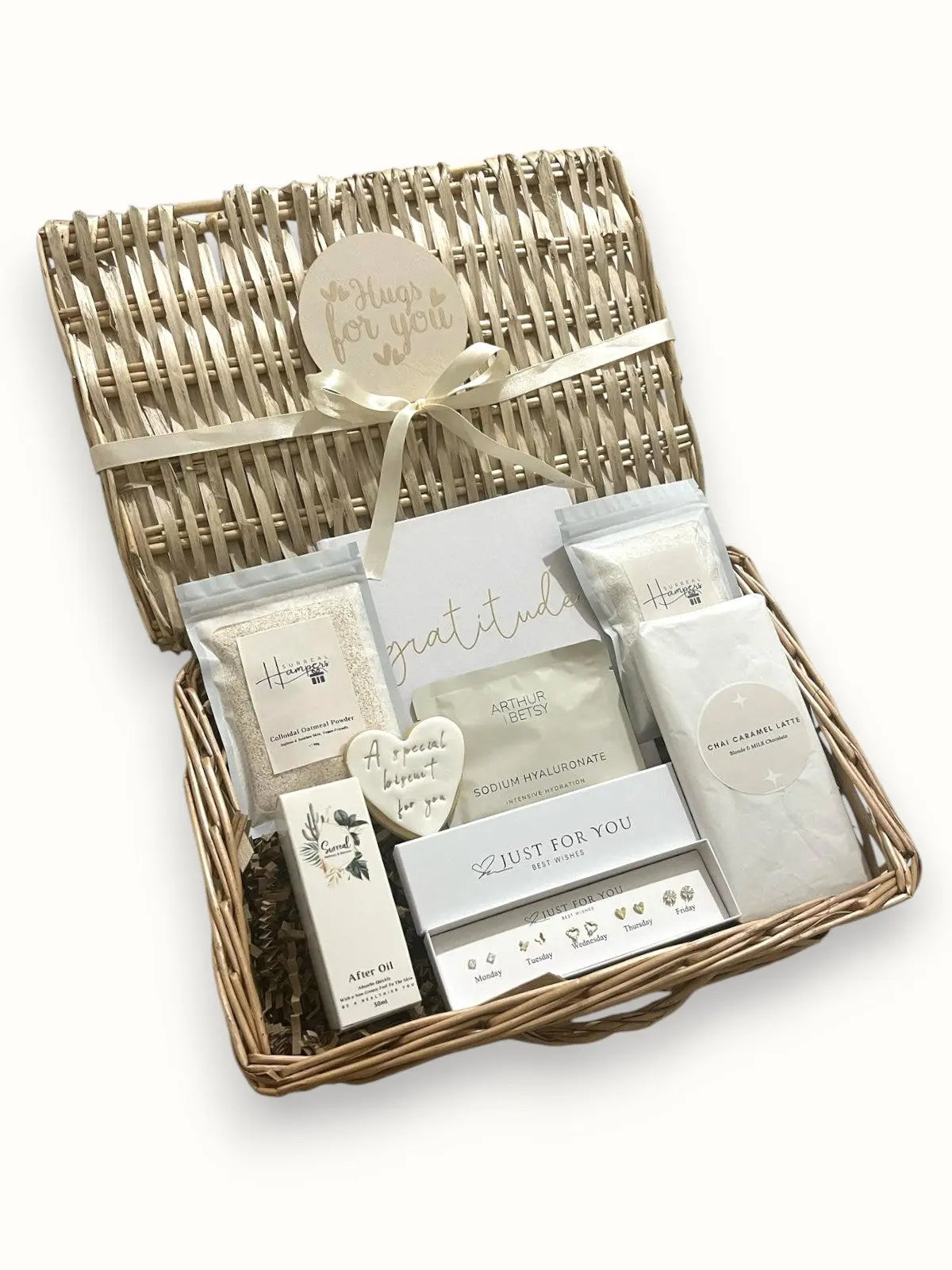 A wicker gift basket containing various items including a sodium hyaluronate face sheet mask, chamomile dreams bath salts, a colloidal oats face mask, five pairs of earrings, a gratitude journal, a 'hugs for you' biscuit, a face mask sheet, a chai caramel latte chocolate bar, and a personalized coaster.