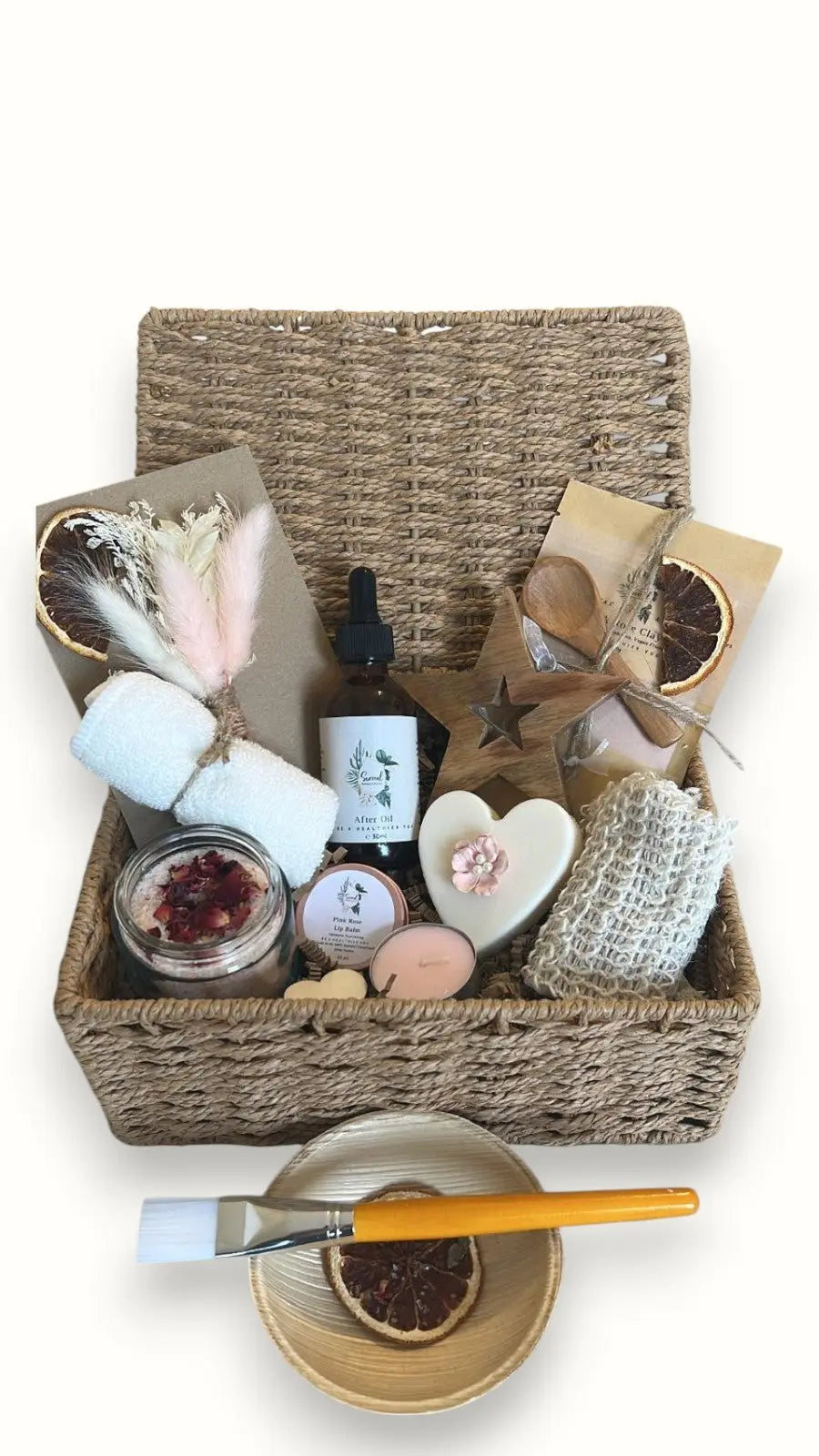 A wicker basket gift set containing various personal care products including a soap, exfoliating soap bag, after oil, clay mask, lip balm, tealight candle, and bath salts. Handmade Hampers 