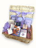 A wicker hamper basket adorned with a purple ribbon, containing various gift items including bath salts, a happy birthday bath bomb, shower steamer, chocolate cookies, a moisturizing hair mist, lavender petals, salt caramel chocolate malt balls, and a personalized coaster.