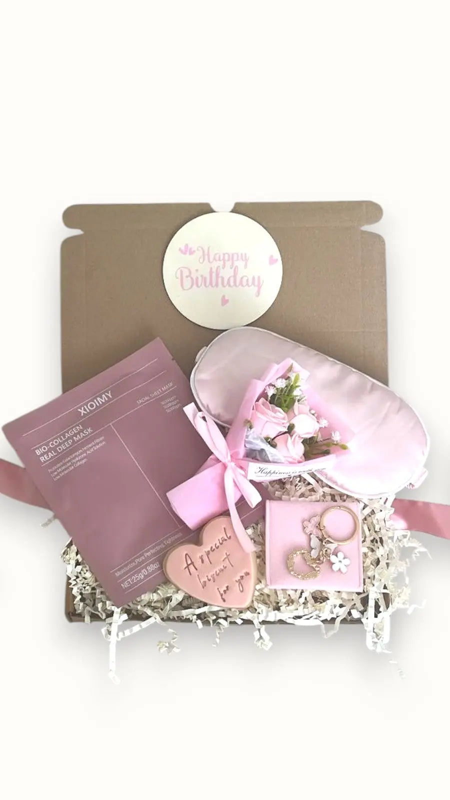 A gift box with a 'Happy Birthday' card, a pink love heart biscuit, a face mask sheet, a butterfly keyring with a bag, a silk sleep mask, and a mini dried flower arranged on a white base.