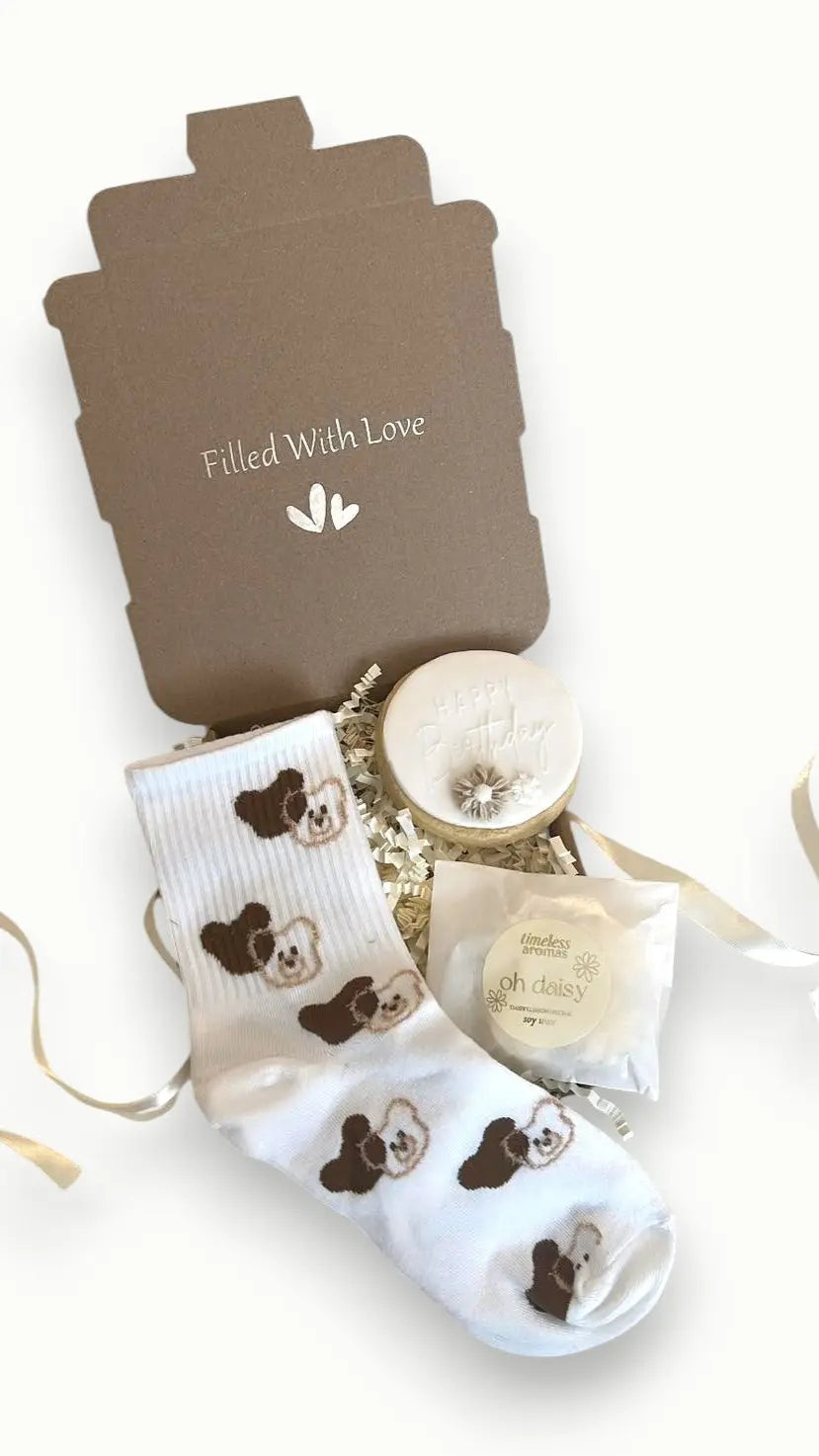 A gift set containing a pair of white bear socks, a 'Happy Birthday' biscuit, and a heart-shaped necklace, all packaged in a box with a 'Filled With Love' card.