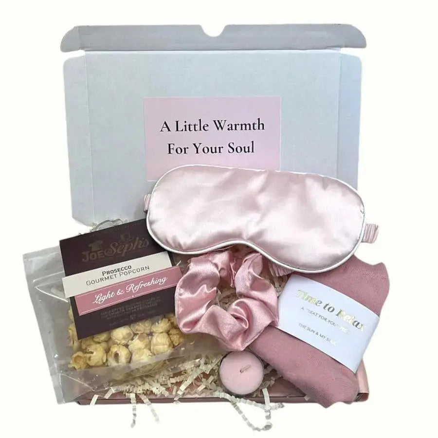 Gift set with pink satin eye mask, pink bath bomb, and chocolate truffles in a box labeled 'A Little Warmth For Your Soul'.