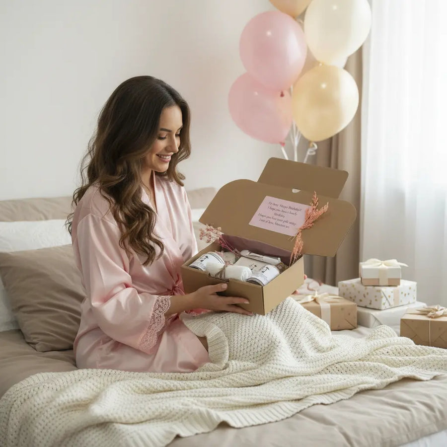 Woman in a pink robe opening a gift box with balloons in the background natural skincare gift box with bath salts, pink clay mask, sugar scrubs