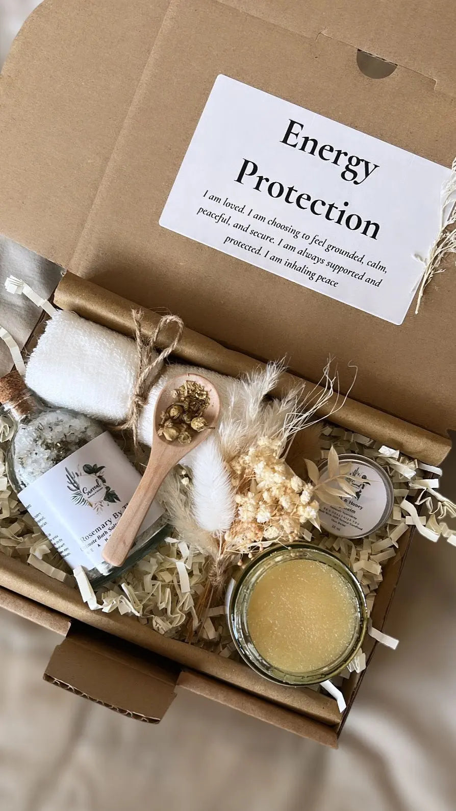 A gift box with various handmade skincare products including a sugar scrub, lip balm, bath salts, a bowl, spoon, and towel, with 'Energy Protection' written on the top cover. Positive affirmation gifts, Christmas gift box and Spa gift set
